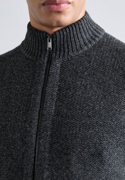 Pull en tricot gris avec un col montant, présentant une fermeture éclair intégrale et un tissu texturé. Le design met en avant un motif subtil et des accents lisses.