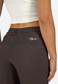 Hnědé kalhoty s hladkým povrchem, mají stahovací pás a dvě zadní kapsy. Na kalhotách je vyšitý malý bílý logo "Pegador".