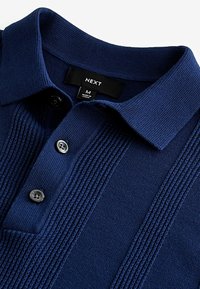 Polo bleu marine avec un motif côtelé texturé sur le devant, patte de boutonnage à trois boutons, et col classique. Étiquette indiquant "NEXT", taille M.