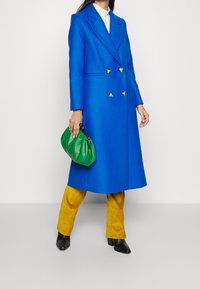 Lungo cappotto blu doppiopetto con bottoni argentati, abbinato a pantaloni gialli in velluto a coste e una borsa verde. Scarpe nere con tacco completano l'outfit.