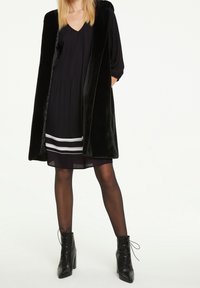 Schwarze Kunstfellweste über einem schwarzen plissierten Kleid mit weißen horizontalen Streifen. Modell trägt schwarze Schnür-Ankle-Boots und transparente schwarze Strumpfhosen.