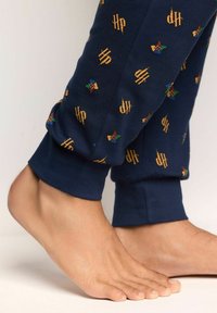 Joggers de algodón azul marino con patrones dispersos en dorado de "HP". Con puños en los tobillos, mostrando pies descalzos y piel suave.