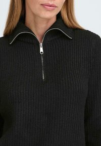 Pulover negru cu textură canelară, cu guler înalt și detalii din fermoar argintiu la gât. Textura pare moale și tricotată.