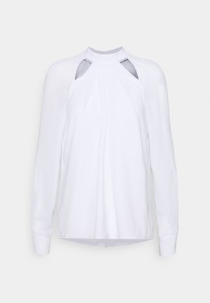 Witte blouse met lange mouwen, hoge halslijn, geplooide voorkant en uitsnijdingen bij de schouders, weergegeven op een effen achtergrond.