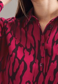 Blusa fucsia con un motivo astratto nero, caratterizzata da un colletto e bottoni. Il tessuto sembra liscio e leggero.