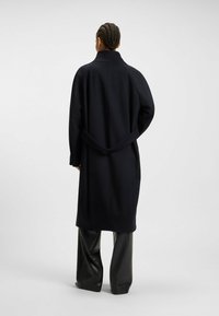 Persona con i capelli intrecciati che indossa un lungo cappotto nero e pantaloni neri a gamba larga, in piedi contro uno sfondo grigio chiaro uniforme.