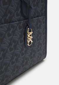 Tote taška s námornickým vzorem, která obsahuje kovový přívěsek s logem ve zlatém provedení. Je vyrobena z odolného materiálu s hladkou texturou a kontrastními akcenty.