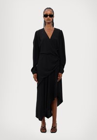 Black wrap dress with long sleeves, asymmetric hem, and soft draping. Features a deep V-neck and smooth texture. Worn with strappy black sandals.