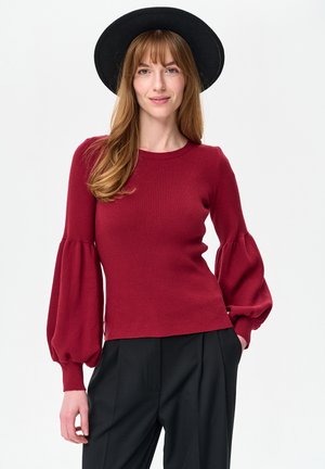 Rode geribbelde trui met lange mouwen en ballonvormige mouwen, gecombineerd met zwarte broek. De stof lijkt zacht met een getextureerde afwerking. Zwarte hoed gedragen.