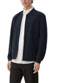 Granatowa kurtka bombowca wykonana z miękkiego materiału, z zapięciem na suwak, delikatnie prążkowanymi mankietami i kieszeniami po bokach, noszona na białej koszuli.