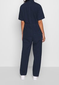 Marinblå bomullsbyxdress med korta ärmar, avslappnad passform, bakficka och elastisk midja. Slät textur och minimalistisk design.