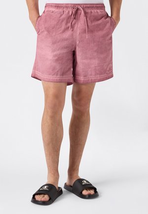 Hombre con pantalones cortos rosa desteñidos con cordón y sandalias negras Champion, de pie con las manos en los bolsillos sobre un fondo blanco.