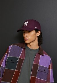 Maroon kapa za bejzbol s bijelim NY logom, nošena s multikoloritnom kariranom flanel košuljom koja sadrži ljubičaste i burgundske nijanse.