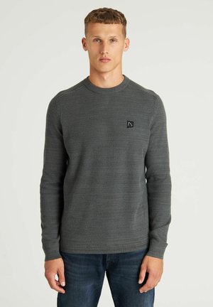 Suéter gris de mangas largas con patrón de punto texturizado, escote redondo y un pequeño parche con logo negro en el pecho, combinado con jeans azules.