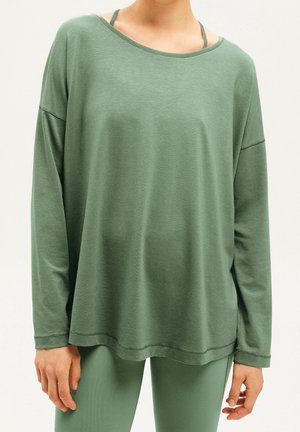 Personne portant un haut ample vert sauge à manches longues et des leggings ajustés assortis, debout devant un fond uni.