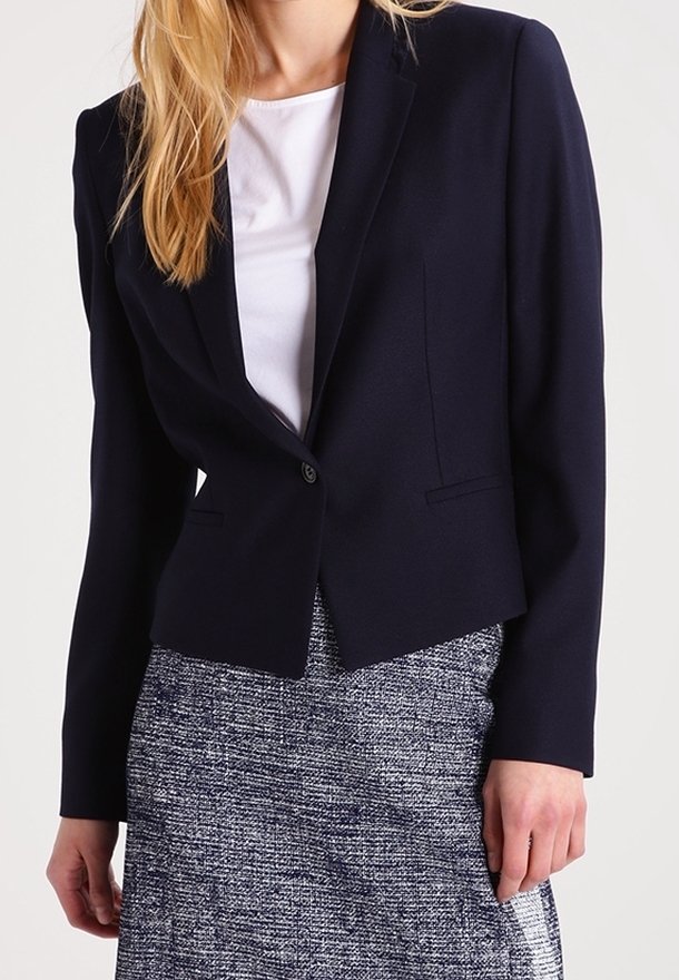 Femme portant un blazer bleu marine ajusté, un haut blanc et une jupe grise texturée, debout avec une main relâchée le long du corps.