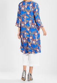 Túnica floral azul con mangas tres cuartos, que presenta un vibrante patrón floral en tonos de rosa y amarillo, llevada sobre pantalones capri blancos.