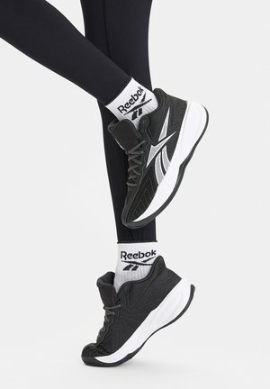 Czarne sportowe buty z białymi akcentami i siateczkową fakturą, w połączeniu z białymi skarpetami Reebok z czarnym logo, noszone z czarnymi legginsami.