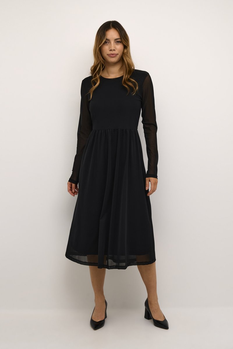 Robe noire à manches longues avec un corsage ajusté et une jupe fluide, en tissu léger ; manches transparentes et encolure ras du cou. Talons noirs.