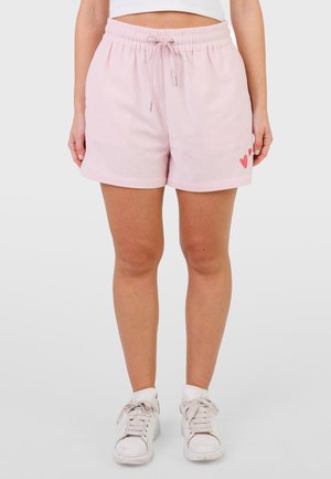 Femme portant un short rose pâle à cordon avec de petits détails en forme de cœur rouge sur le côté gauche, des baskets blanches et un top court blanc.