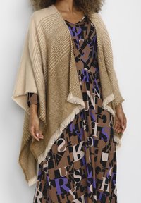 Beige vävd poncho med fiskbensmönster, fransade kanter, lager-på-lager över en brun klänning med djärva, blockbokstäver i lila och svart.