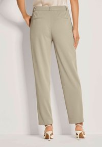 Vrouw in beige op maat gemaakte broeken en witte hoge hakken, staande met haar rug naar de camera.