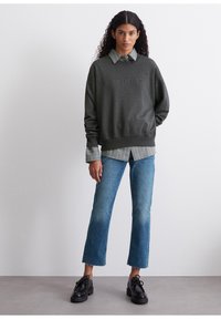 Felpa grigia scura con testo in rilievo, indossata sopra una camicia a righe chiara con colletto. Abbinata a jeans blu e scarpe nere con lacci.