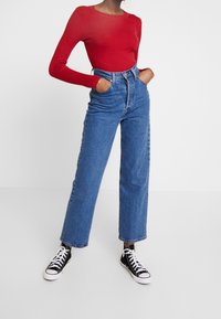 Maglietta rossa a maniche lunghe a coste abbinata a jeans blu a vita alta. I jeans presentano una silhouette dritta, e le sneakers nere alte completano il look.