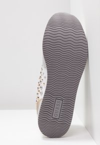 Sohle eines Guess-Sneakers mit wellenförmigem Profil, dekorativen metallischen Nieten an der Seite und glitzerndem Fersen-Detail vor weißem Hintergrund.