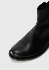 Bottine noire avec un dessus en cuir lisse, bout rond et fermeture éclair à l'arrière. Dotée d'une semelle en caoutchouc plate et basse.
