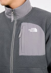 Giacca in fleece grigio caratterizzata da un colletto alto, chiusura con zip e una tasca sul petto con una patta grigia chiara. Logo stampato sulla tasca.