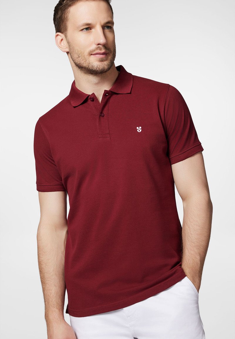 Burgundowa koszulka polo z bawełny z klasycznym kołnierzykiem, zapięciem na dwa guziki i małym białym logo na lewej piersi. Krótkie rękawy.