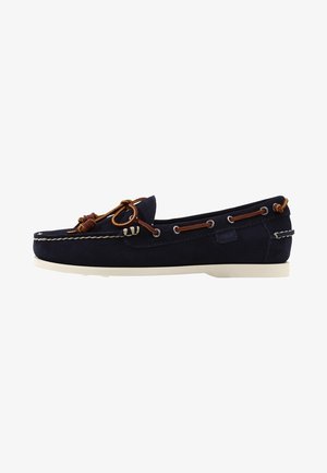Chaussure bateau en daim bleu marine avec coutures blanches, lacet en cuir marron noué en nœud et semelle en caoutchouc blanche.