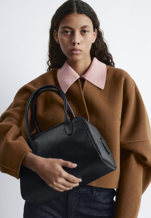 Black leather handbag with structured design, rounded handles, and a textured finish. The model wears a brown sweater with a pink collar.