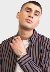 Cămașă cu dungi în nuanțe de gri, portocaliu și bleumarin; are un guler tip spread și mâneci lungi. Pur tată peste un tricou alb. Brățări și inele din argint.