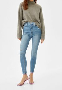 Jeans ajustados de tiro alto en denim azul suave, con un desgarro en la rodilla izquierda. Combinados con un suéter de punto gris y sandalias de tacón rosa.