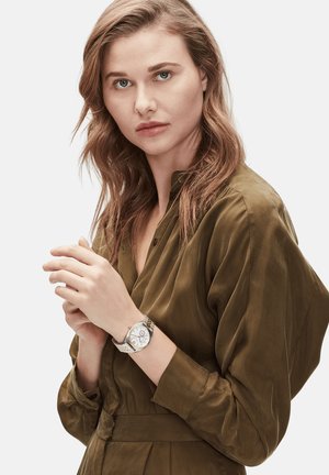 Silberne Armbanduhr mit einem runden weißen Zifferblatt und silbernem Armband. Das Modell trägt ein strukturiertes olivgrünes Hemdkleid mit einem geknöpften Kragen und taillierter Taille.