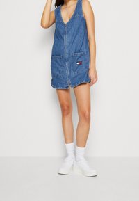 Kobieta w bezrękawnikowej, niebieskiej sukience dżinsowej z zamek błyskawicznym z przodu i kieszonką na łatkę, w połączeniu z białymi skarpetkami i platformami sneakersami.