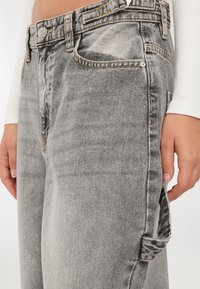 Osoba w wyblakłych szarych dżinsach i białej bluzce z długim rękawem, z rękami swobodnie opuszczonymi wzdłuż ciała.