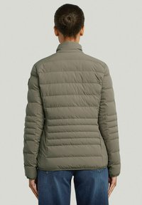 Olijfgroen pufferjack met verticale doorgestikte patroon, staande kraag en lange mouwen. Draag het over blauwe jeans, wat een losse pasvorm toont.