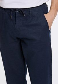Námorně modré, lehké bavlněné kalhoty s páskem na stažení, předními kapsami a hladkou texturou, částečně viditelné v pase a na stehnu.