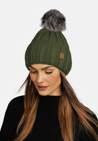 Knitted green beanie with ribbed texture and a faux fur pom-pom on top. Features a small brown leather tag on the side.