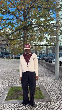 Tejszínszínű kardigán bordázott mintával, alatta bordó ing, sötét, széles szárú nadrág, valamint bordó sapka fehér felirattal és logóval. Kint áll a szabadban.