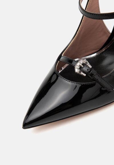 Zapato de charol negro de tacón alto con punta afilada, correa ajustable con hebilla decorativa de plata, textura suave y diseño minimalista.