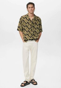 Chemise à manches courtes en vert olive avec un motif géométrique noir, associée à un pantalon crème et des sandales noires.