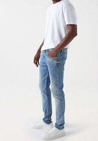 White short-sleeve t-shirt, light blue jeans, slim fit, faded detailing, and white sneakers, standing against a neutral background.
