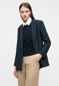 Γυναίκα που φοράει μπλε σκούρο σακάκι πάνω από μπλε σκούρο πουλόβερ και λευκό πουκάμισο με γιακά, με μπεζ παντελόνι προσαρμοσμένο, ποζάρει μπροστά σε απλό ανοιχτό φόντο.