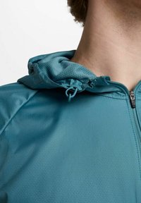 Chaqueta con cremallera en color verde azulado, con una superficie texturizada, que cuenta con una capucha ajustable y un sutil patrón geométrico en la tela.
