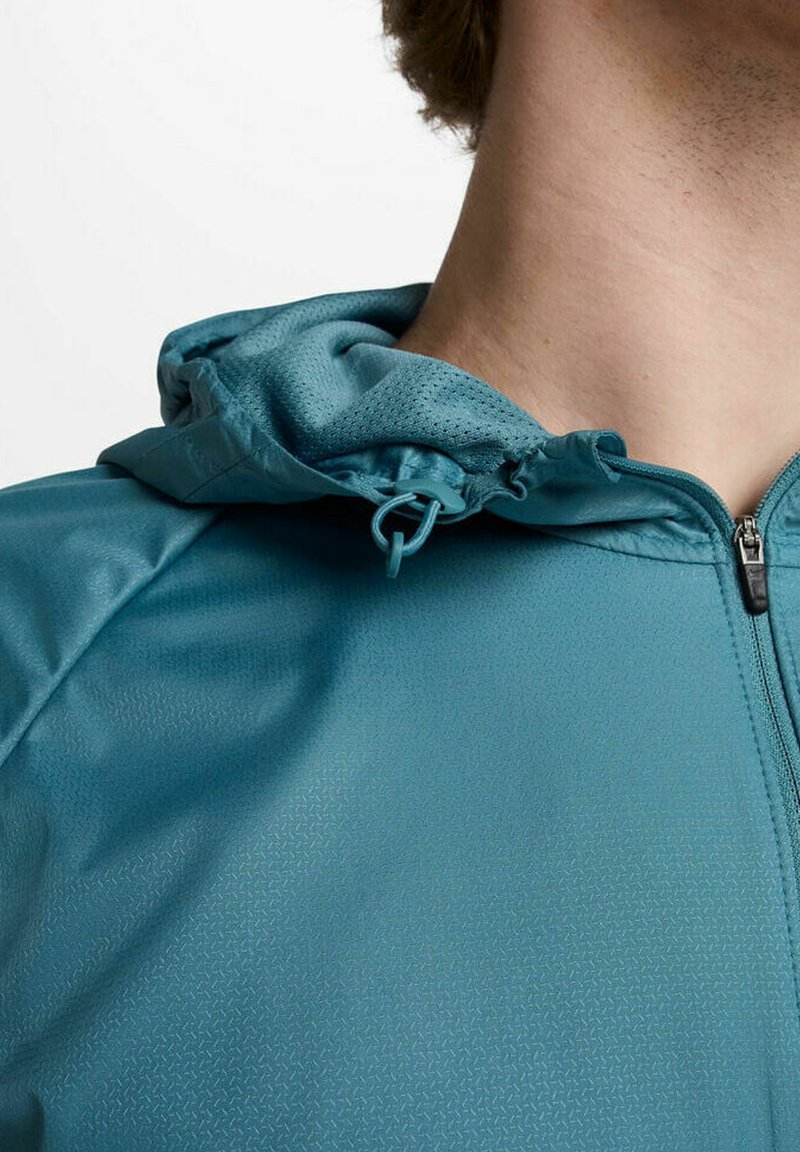 Veste zippée teal avec une surface texturée, dotée d'une capuche à cordon de serrage et d'un motif géométrique subtil sur le tissu.