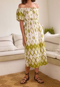 Robe à épaules dénudées en motif ikat blanc et vert, avec une taille élastique et des manches courtes bouffantes. Des sandales noires à brides complètent le look.